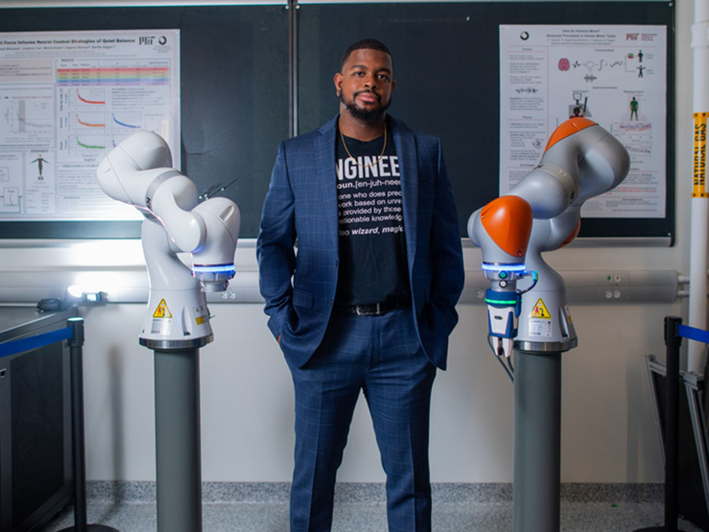 Michael West, wearing a suit with an "Engineer" T-shirt, stands in front of chalkboard, flanked by two robotic arms.