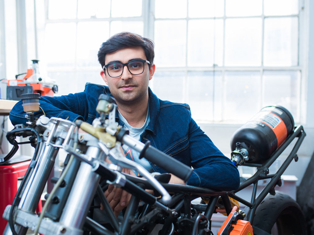 Adi Mehrotra smiles while resting his arms on a handmade motorcycle. Mehrotra is inside a shop and windows in background.