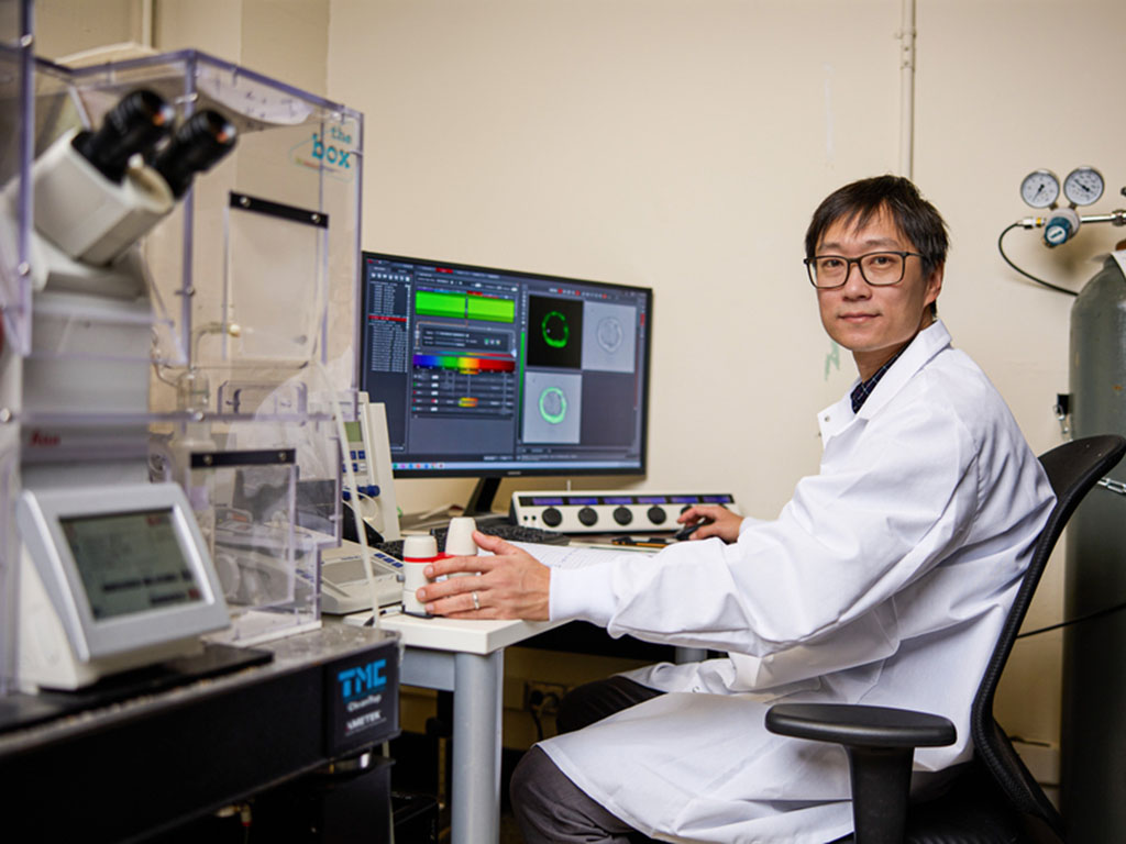 Guo, wearing a lab coat, sits at a desk in front of a computer inside a lab with large gas canisters and microscopes around him. The computer screen shows green and rainbow bars, and neon green circles.