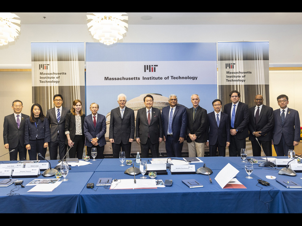 The 13 participants in the roundtable discussion stand side by side. In front of them is a wide table strewn with microphones, notepads, name cards, and water glasses. Behind them is a screen showing a photo of MIT’s Great Dome, with the MIT logo and the 