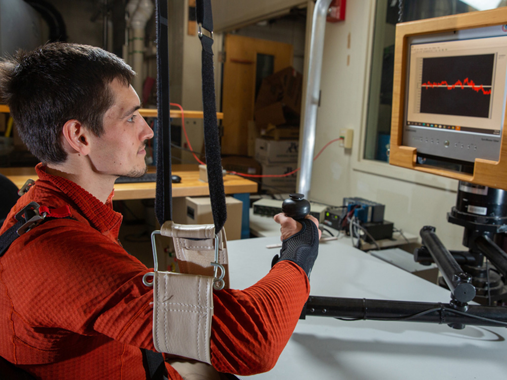 Hermus looks at a computer display while his arm is in a sling. He grips a joystick while wearing a black wrist strap.